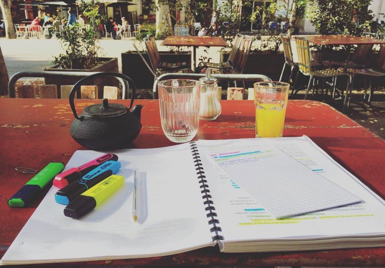 Un cahier de cours surligné, des feutres et un thé, sur une terrasse au soleil