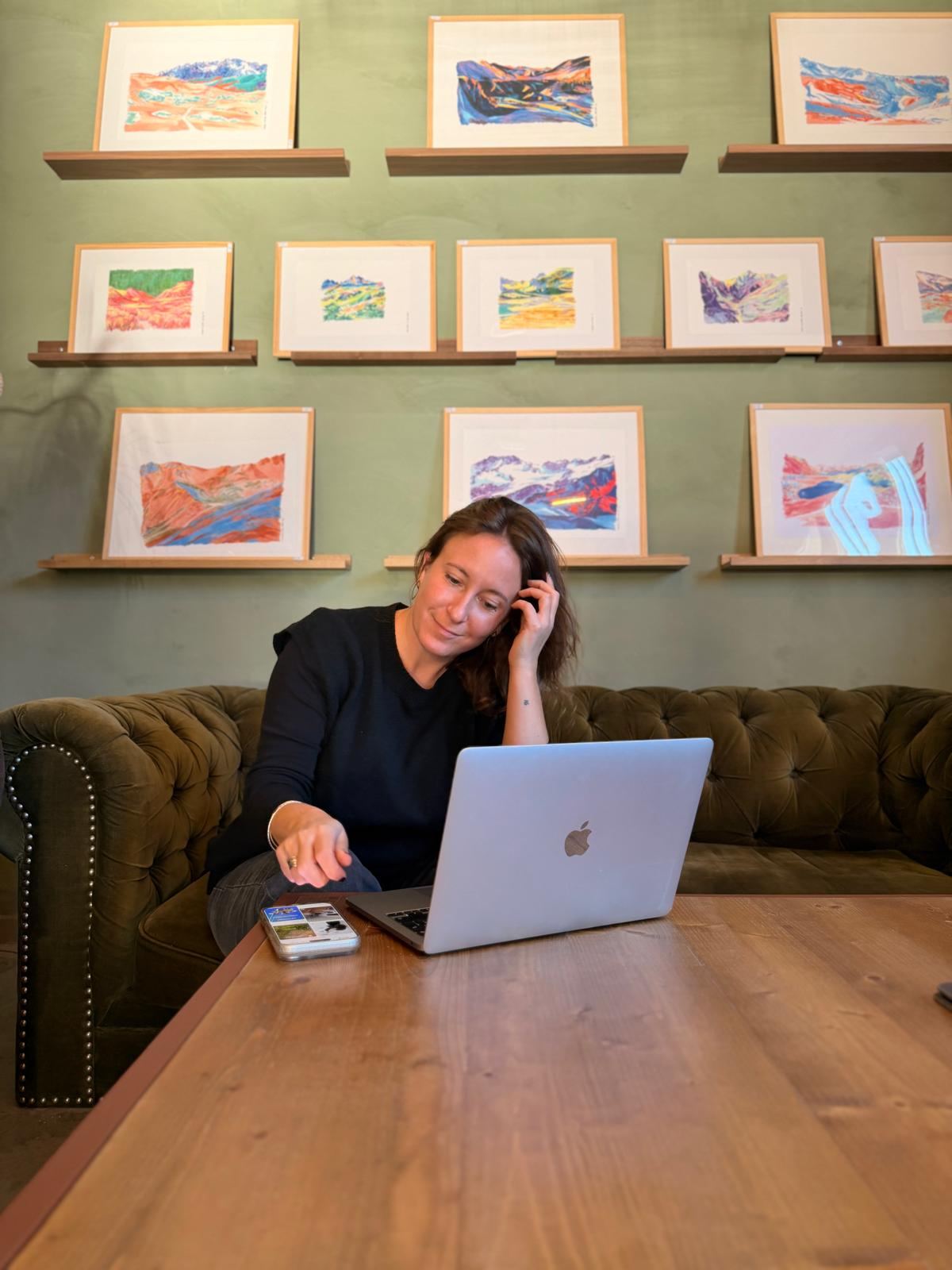 Laure Gasquet sur un canapé en velours vert, devant son ordinateur, sous un mur de tableaux colorés