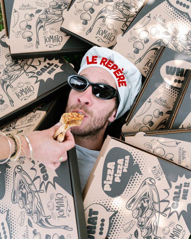 Romain, lunettes noires et toque du Repère, croque une part de pizza au milieu des boîtes
