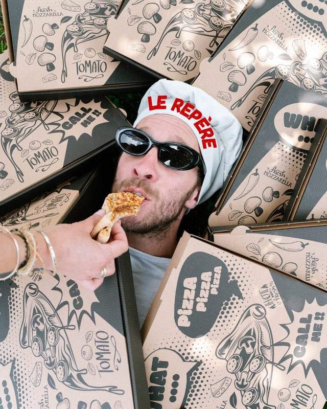 Romain, lunettes noires et toque du Repère, croque une part de pizza au milieu des boîtes
