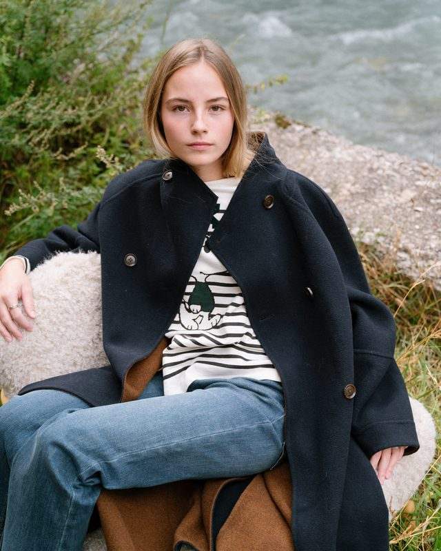 Une jeune femme en manteau noir et pull rayé pose assise dans l’herbe