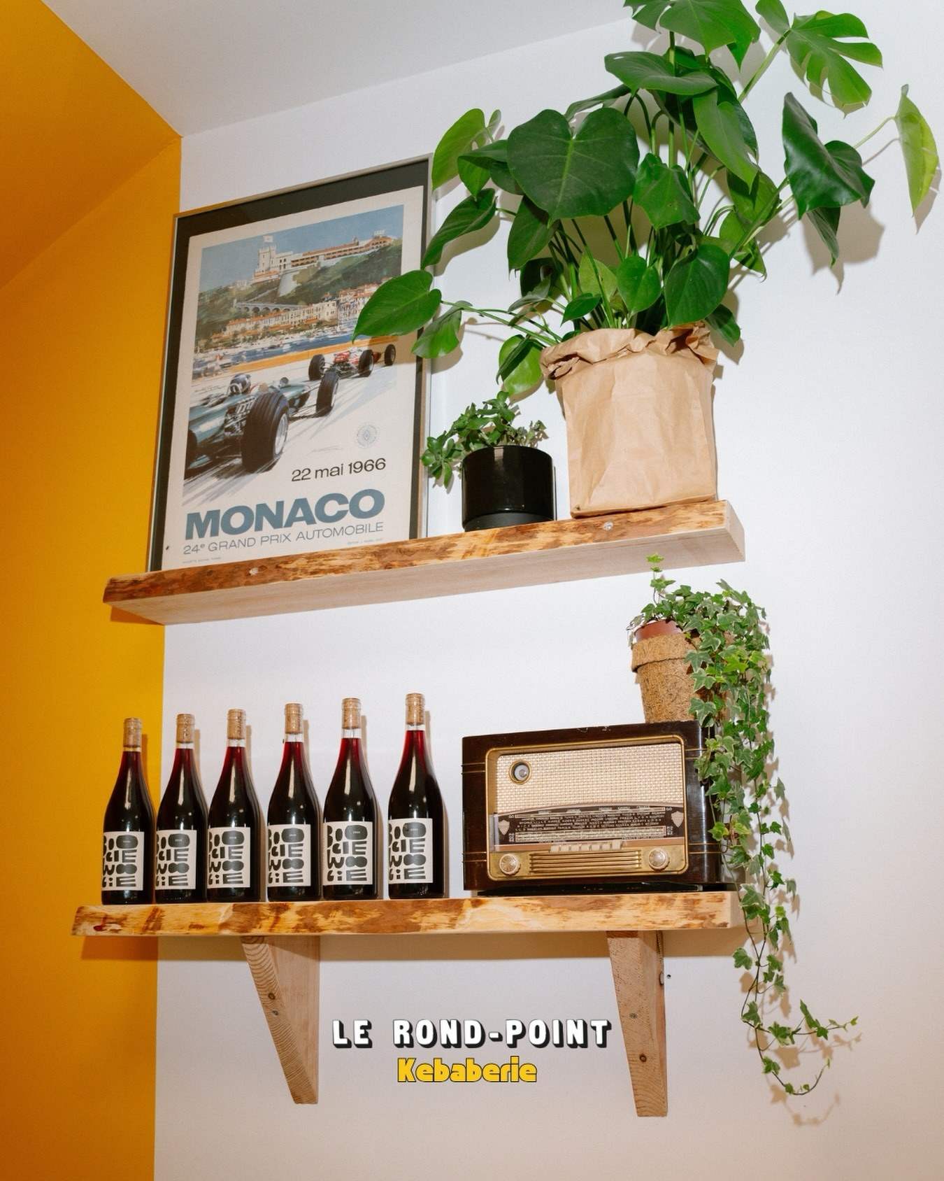 Une étagère en bois sur un mur jaune : affiche de Monaco, plantes, bouteilles de vin et vieille radio du Rond-Point
