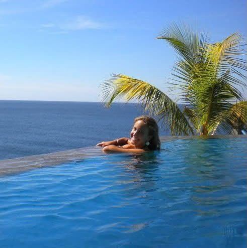 Laure dans une piscine à débordement face à la mer, sous un palmier